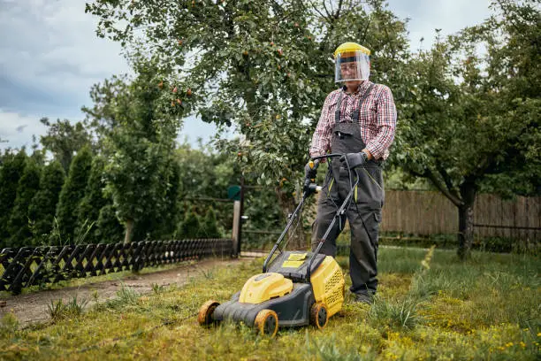 Photo of Active senior man in overalls and with protection mowing the grass in his backyard with push mower