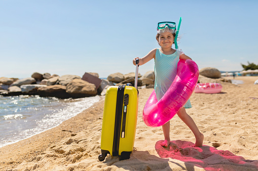 Excited little 4 year old girl on a beach wearing scuba mask and inflatable ring, while holding suitcase