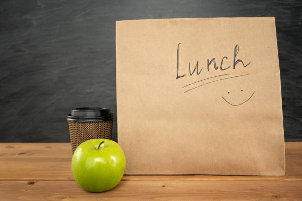 экологически чистый коричневый бумажный обеденный пакет на деревянном столе с яблоком и кофейной чашкой. меловая доска на заднем плане. ве� - packed lunch paper bag apple lunch стоковые фото и изображения