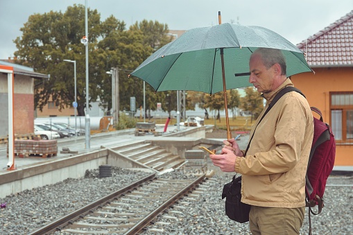 Portrait of a mature traveller texting on smartphone with an umbrella standing on the platform of a train station. Unfinished railway line. When the train arrives.. Funny concept of futile situation.