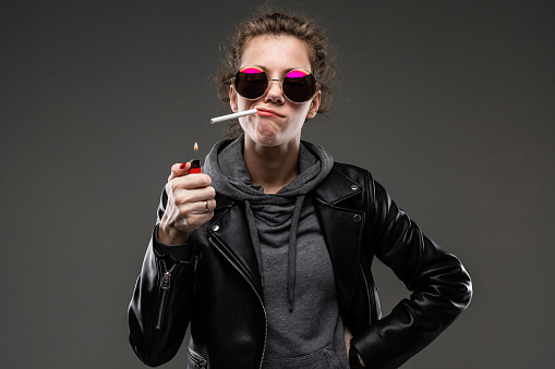 Caucasian girl with rough facial features in a black jacket lights a cigarette isolated on black background.