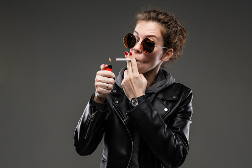 Caucasian girl with rough facial features in a black jacket lights a cigarette isolated on black background.