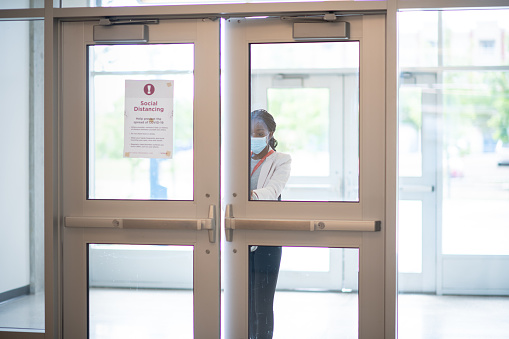 A woman opens the door of a large, sterile building. She is wearing a mask and business clothing.