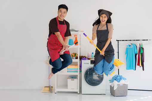 asian young couple having fun doing houseworks together. hasband and wife enjoy housecleaning riding on mop and jump like witches broom