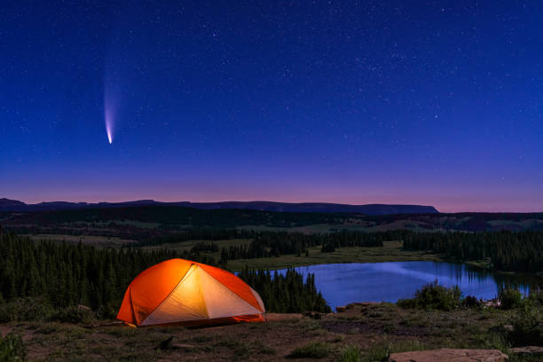 Tent Lit Up with NEOWISE Comet in Sky - Landscape scenic view with comet in sky at dawn and orange glowing tent overlooking scenic lake and mountain wilderness backcountry setting.