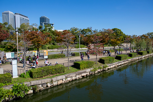 The grounds and pedestrian walkway of the Osaka Castle n clear autumn day, as seen from Osaka Castle.  People walk through the gardens enjoying the scenics.