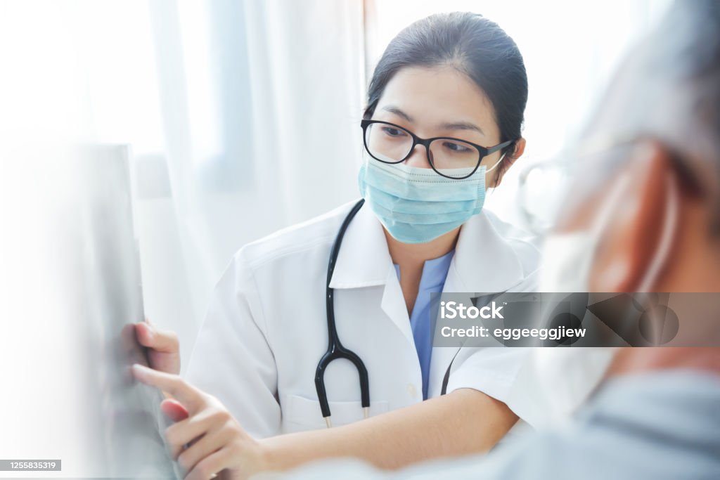 Asian woman Doctor in surgical mask giving advice to old man patient Physician talking with senior man at clinic pandemic during Covid-19 or Coronavirus. Asian woman Doctor wear eyeglasses and surgical mask giving advice to Elderly patient in medical room at hospital Doctor Stock Photo