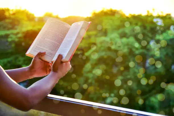 © 2017 Kosal Hor All Rights ReservedClose-up of man's hands holding a book by the window for reading against sunset with beautiful nature background. warm tone and soft focus.