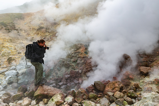Male photographer takes pictures of aggressive volcanic landscape, hot spring, eruption fumarole, gas-steam activity in crater active volcano. Kamchatka Peninsula, Russian Far East - September 7, 2014