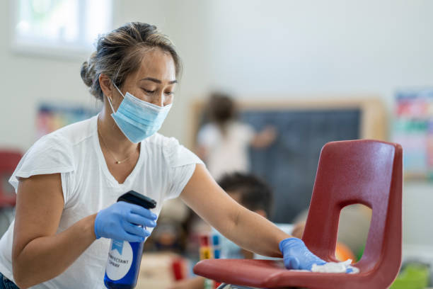 Daycare instructor using a disinfectant to clean classroom equipment Facilitator at a daycare cleaning the chair between use while children play in the background to protect from the transfer of germs during COVID-19 school cleaning stock pictures, royalty-free photos & images