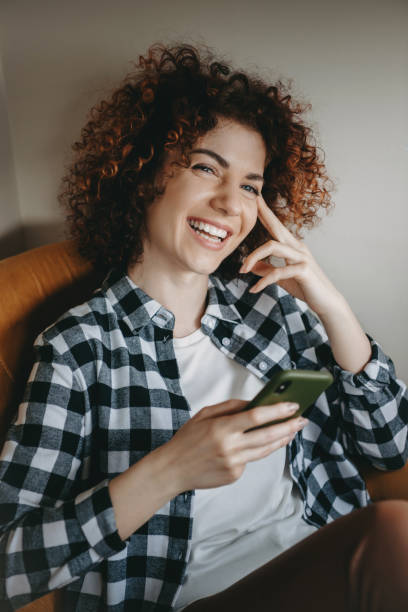 istock Cute caucasian woman with curly hair smiling while sitting on the sofa and chatting on mobile 1255845310
