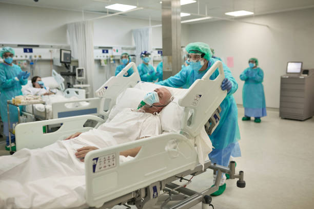 Healthcare Worker Moving COVID-19 Patient in Hospital Bed Member of medical team in full protective workwear pushing senior Caucasian male patient in hospital bed past camera. hospital room stock pictures, royalty-free photos & images