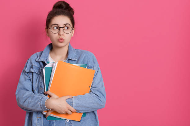 istock Thoughtful pretty woman looks pensively aside with rounded lips, holds paper folder, has pleasant appearance, being deep in thoughts. Copy space for advertisement. 1255857867