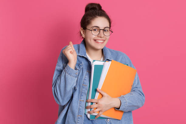 istock Young smiling student in eyeglasses and denim jacket standing with paper folders isolated over rosy background, clenching fist, looking smiling at camera. 1255857870