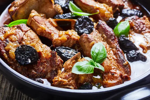 Pork ribs tajine stew with prunes, red wine, broth, onion, and garlic, decorated with fresh basil, served on a black baking dish on rustic wooden background with a linen towel, close-up