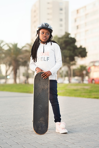 Portrait of a young african girl wearing helmet standing with a skateboard outdoors and looking away