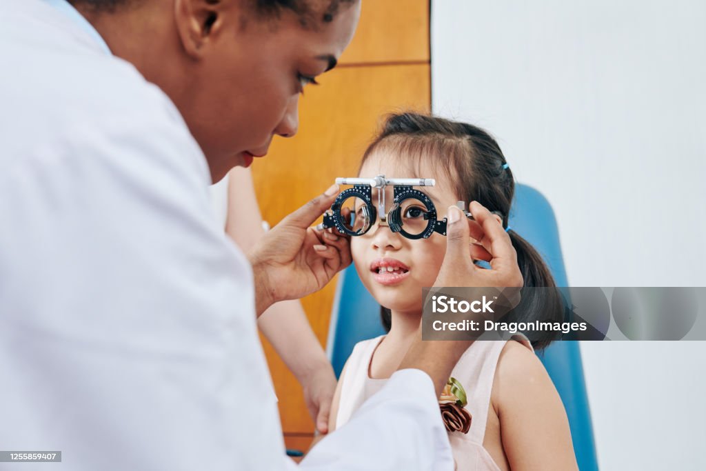 Girl visiting pediatric optometrist Pediatric optometrist helping little girl to put on test glasses when checking her eyesight Child Stock Photo