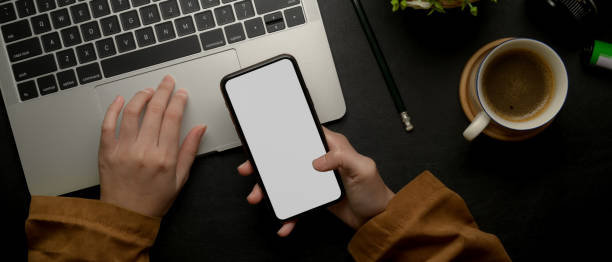 istock Female freelancer using mock-up smartphone while working with laptop on dark office desk 1255861386