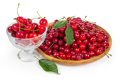 Freshly picked sour cherries on the wooden dish and sweet cherries in glass cup, leaves of cherry tree on a white background