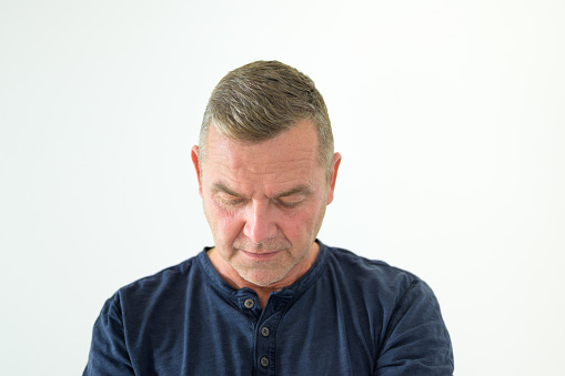 Depressed middle-aged man in a casual blue top looking down with sad serious expression in a head and shoulders portrait on a white background