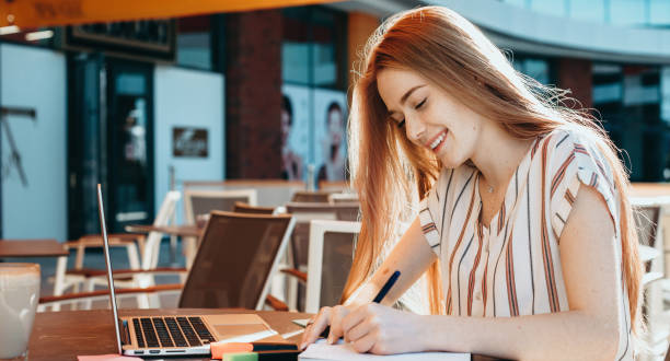 istock Red haired caucasian student with freckles doing homework at the computer in a cafeteria writing a in book and smile 1255869320
