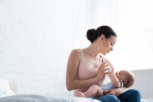 beautiful woman feeding cute infant son while holding baby bottle