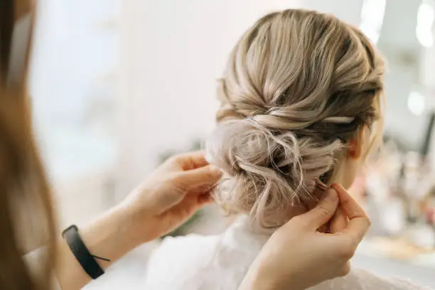 Close-up of hands of hairdresser making hairstyle unrecognizable young blonde woman in beauty salon, back view. Concept of beauty, fashion and stylish makeup.