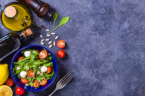 Top view of a blue bowl full of fresh salad surrounded by various ingredients like arugula leaves, cherry tomatoes, pine nuts, and some salad dressing ingredients like balsamic Vinegar, lime, olive oil and pepper. All the objects are at the left side of the image leaving a useful copy space at the right side on a rustic grey bluish backdrop. Studio shot taken with Canon EOS 6D Mark II and Canon EF 24-105 mm f/4L