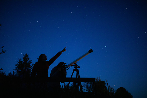 Mother observing stars on the night sky with daughter standing on the viewpoint. Daughter looking stars through the telescope and the mother points with finger in which direction should looking.