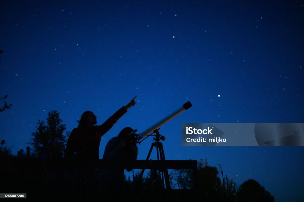Mother and daughter are watching stars Mother observing stars on the night sky with daughter standing on the viewpoint. Daughter looking stars through the telescope and the mother points with finger in which direction should looking. Astronomy Stock Photo
