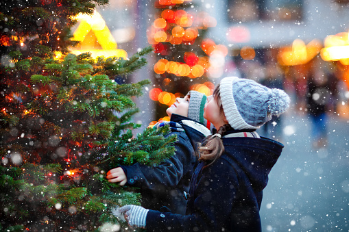 Two little kids, boy and girl having fun on traditional Christmas market during strong snowfall. Happy children enjoying traditional family market in Germany. Twins standing by illuminated xmas tree