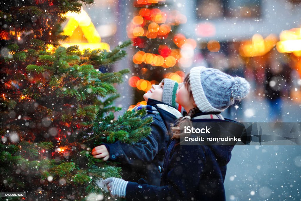 Two little kids, boy and girl having fun on traditional Christmas market during strong snowfall. Happy children enjoying traditional family market in Germany. Twins standing by illuminated xmas tree. Two little kids, boy and girl having fun on traditional Christmas market during strong snowfall. Happy children enjoying traditional family market in Germany. Twins standing by illuminated xmas tree Christmas Stock Photo