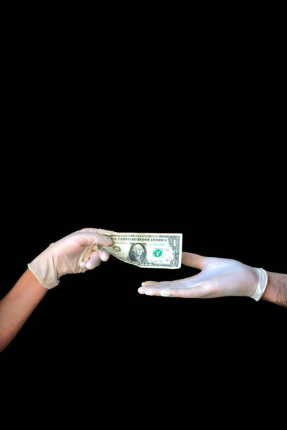 istock Two hands with medical gloves paying with a one Dollar bill on black background. 1255891685
