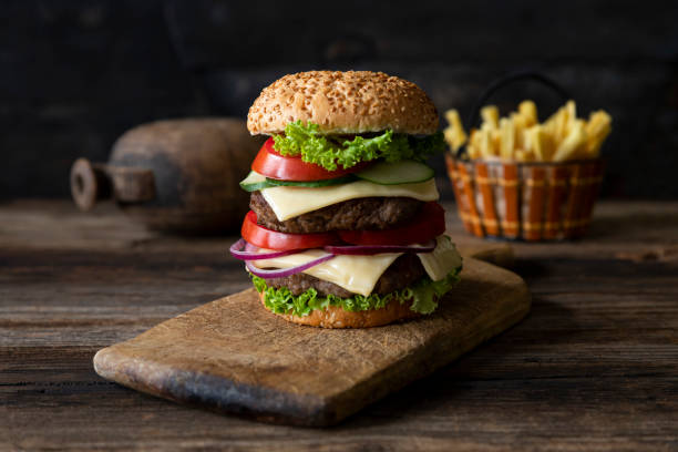 istock Hamburger with tomatoes, onions, cucumber, lettuce and melting cheese served on a  rustic wooden board with french fries and vegetables in background 1255895681
