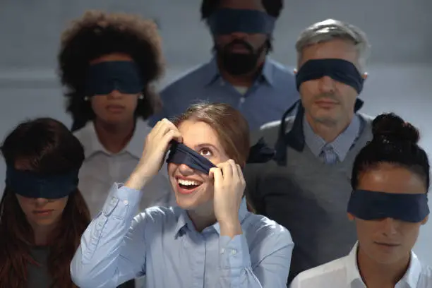Group of blindfolded business people in the dark in the office while one of them is happy to finally see the reality.
