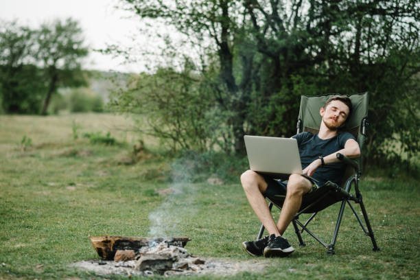 istock Young freelancer relaxing in forest. Man working on laptop on nature. Remote work, outdoor activity in summer. Travel, hiking, technology, tourism, people concept - man sitting on chair outdoors. 1255901496