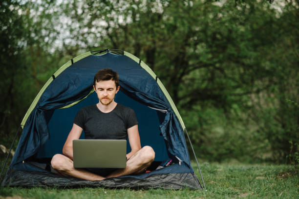 man working on laptop in tent in nature. young freelancer sitting in camp. relaxing in camping site in forest, meadow. remote work, outdoor activity in summer. serious male relaxing, work on vacation. - travel ipad isolated backpack imagens e fotografias de stock