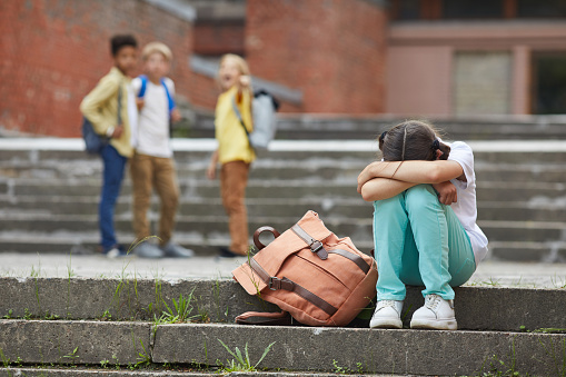 Full length portrait of crying schoolgirl sitting on stairs outdoors with group of teasing children bullying her in background, copy space