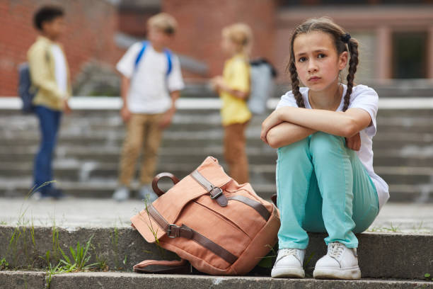 School Outcast Full length portrait of sad schoolgirl looking at camera while sitting on stairs outdoors with group of children in background, copy space school exclusion stock pictures, royalty-free photos & images