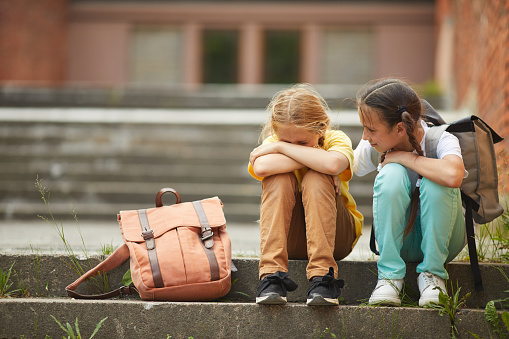 Full length portrait of teenage schoolgirl crying while sitting on stairs outdoors with smiling friend comforting her, copy space