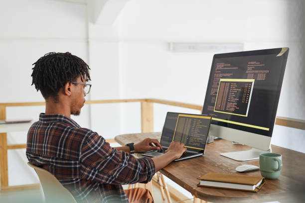 African Computer Programmer Writing Code African-American IT developer typing on keyboard with black and orange programming code on computer screen and laptop in contemporary office interior, copy space website design stock pictures, royalty-free photos & images