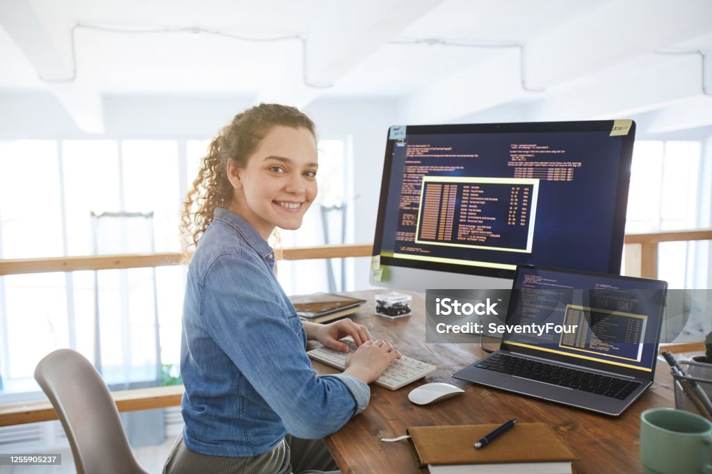 Female IT Developer at Workplace Portrait of female IT developer smiling at camera while typing on keyboard with black and orange programming code on computer screen and laptop in contemporary office interior, copy space Computer Programmer Stock Photo