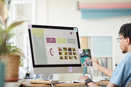 Portrait of young African-American photographer managing photo stock marketplace while using computer at desk in home office with photo website on screen, copy space