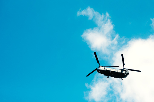A large Chinook helicopter passes directly above, with blue sky and white clouds in the background