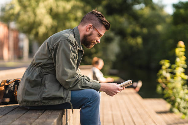 człowiek siedzący w parku czytając książkę - men reading outdoors book zdjęcia i obrazy z banku zdjęć