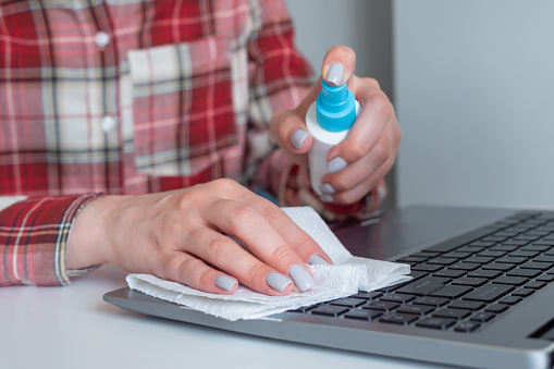 Woman hands spraying antiseptic, cleaning laptop keyboard with disinfectant wet wipe on white table. Disinfection, protection, prevention, housework, COVID 19, coronavirus, safety, sanitation concept