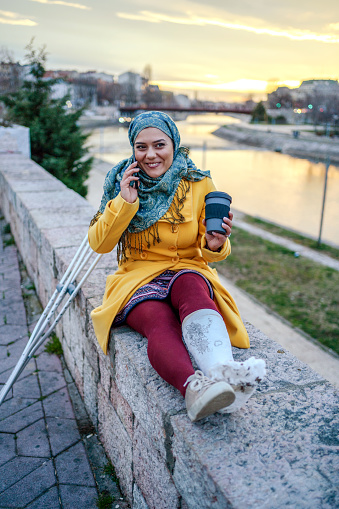 Young beautiful smiling Muslim woman with headscarf talking on smartphone and sitting on quay.