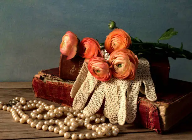 Orange Ranunculus Flowers on old vintage books on wood table with vintage lace gloves, pearls with a dark turquoise blue background.