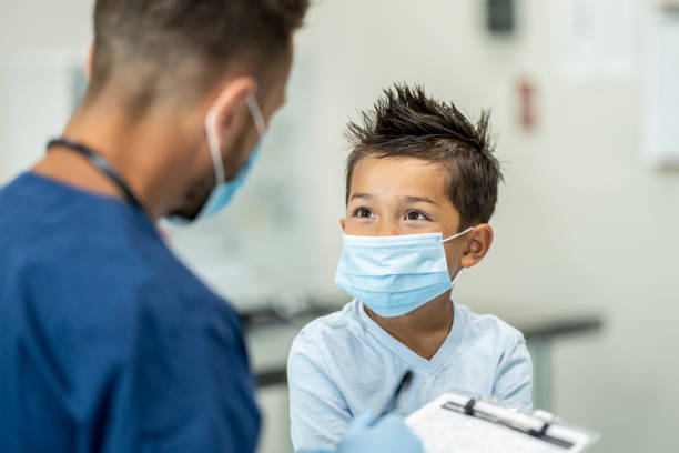 Young boy wearing a mask at a doctors appointment Paediatrician doctor examining a child while wearing a protective face mask in a clinic setting to protect from the transfer of germs during COVID-19. coughing photos stock pictures, royalty-free photos & images
