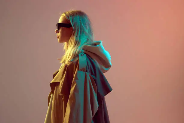 Portrait of a cool stylish young girl, posing in hoodie, sunglasses and cloak on a neon background. High quality photo